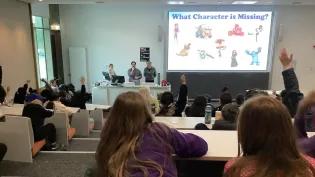 lecture theatre seen from back row, full of primary school children, many with hands raised to answer the question displayed on the lecture theatre screen with three lecturers stood beside lectern