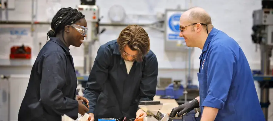 Two engineering students and their supervisor in a mechanical engineering workshop wearing lab coats and safety glasses