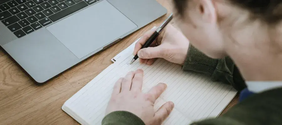 Person writing in a blank notebook, with an open laptop nearby