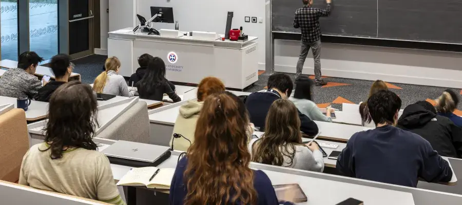 Students sitting in a lecture theatre, with a lecturer writing on the blackboard.