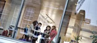 Students in discussion around a table, photo taken from outside building, looking through large glass windows