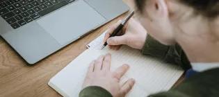 Person writing in a blank notebook, with an open laptop nearby