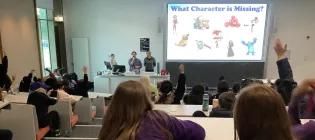 lecture theatre seen from back row, full of primary school children, many with hands raised to answer the question displayed on the lecture theatre screen with three lecturers stood beside lectern