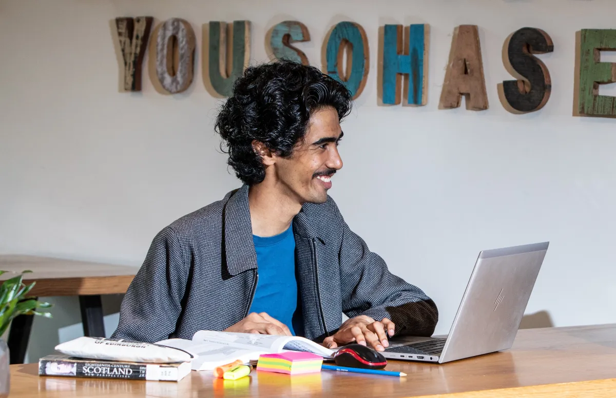 a smiling student sitting at a desk with laptop and books || Undergraduate online interactive talks