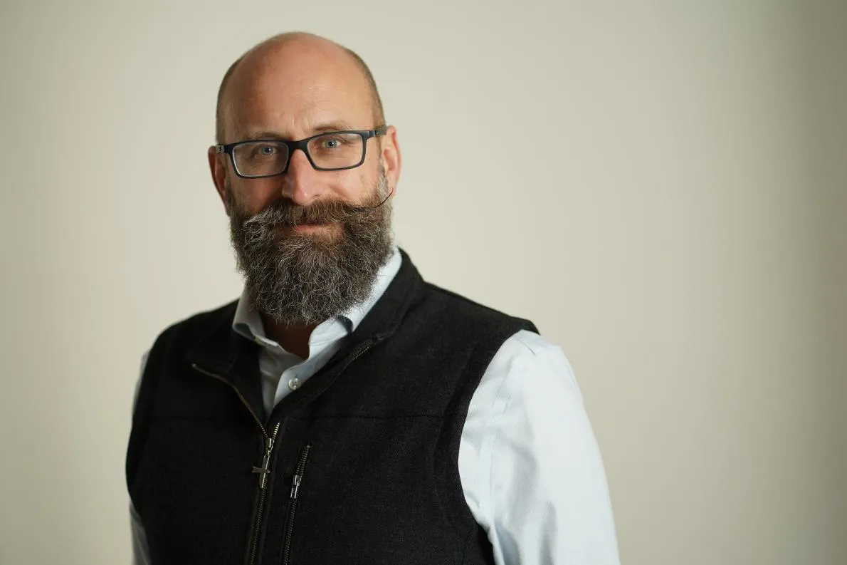 Middle-aged man, bald, brown beard, handle bar moustache, glasses, wearing black vest over white shirt