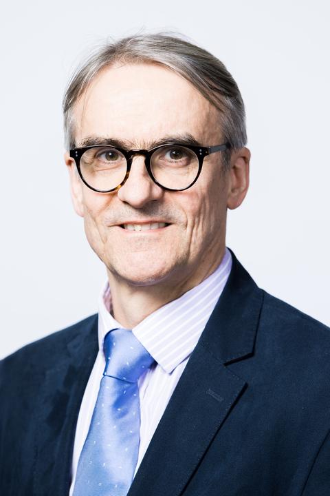 Headshot of Prof Markus Mueller wearing navy suit jacket, white shirt and blue tie