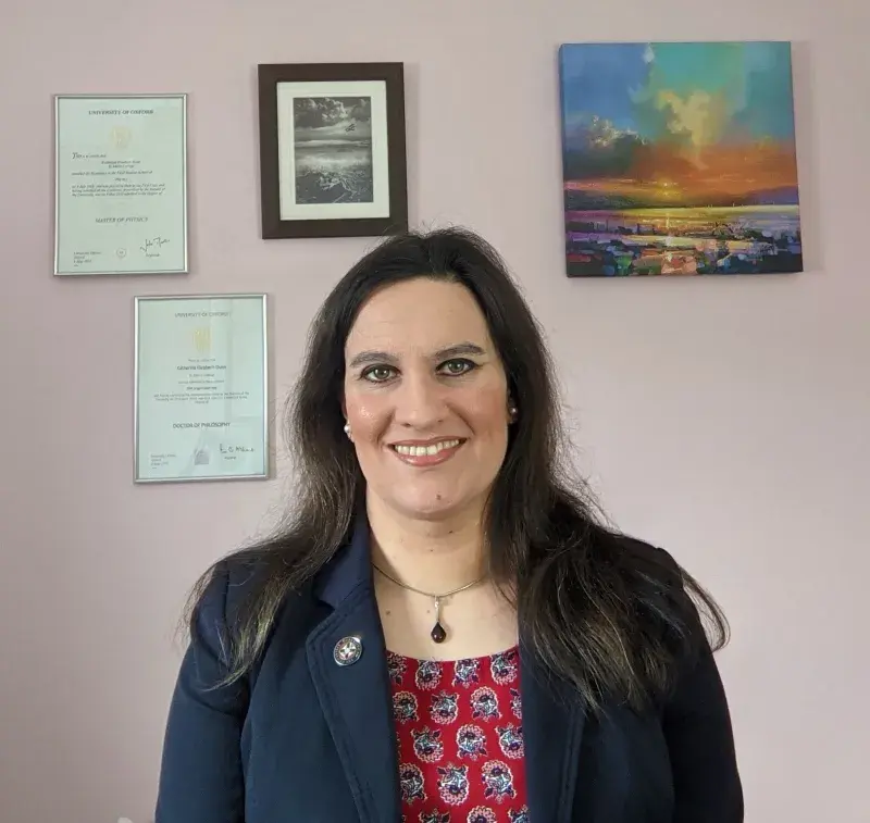Katherine Dunn portrait photo wearing blue jacket and red top with pictures and certificates hanging on wall behind