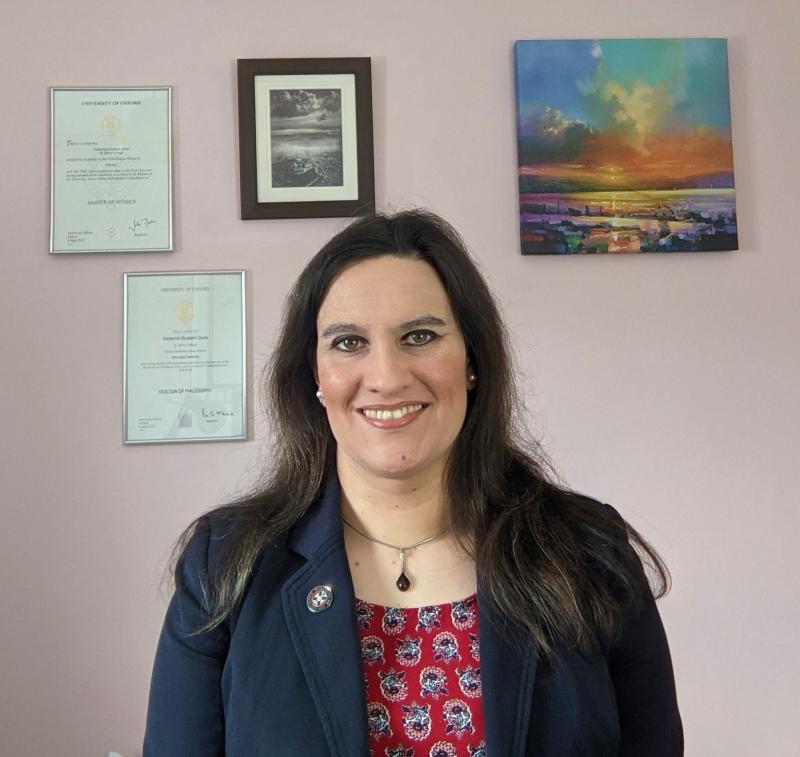 Katherine Dunn portrait photo wearing blue jacket and red top with pictures and certificates hanging on wall behind