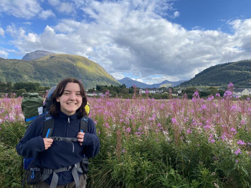 Student hiking through a meadow of wild flowers with hills and mountains behind her