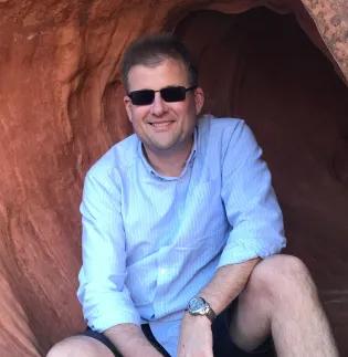 Alex Radford smiling, wearing shades, sitting in a red sandstone cave wearing a light blue shirt and dark shorts shorts